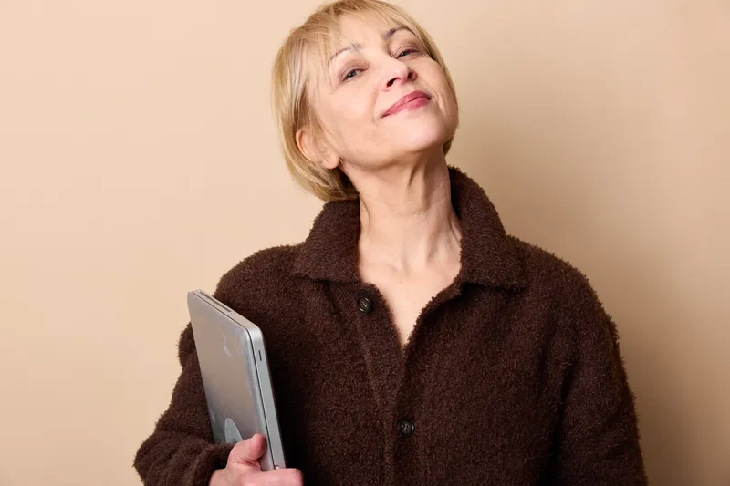Elderly woman, smartphone, communication, portrait, studio beige background, smiling with eyes closed, holding phone in hands, warm calm mood, casual coat, reading message softly