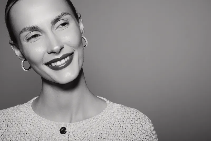 Smiling caucasian woman with light skin and short hair wearing earrings and knit sweater looks away on grey background.