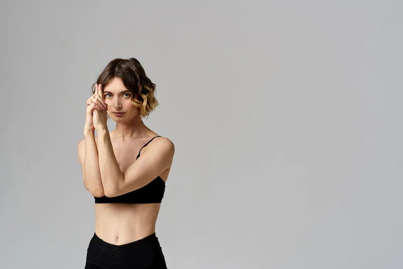 Стройная женщина в черных леггинсах на сером фоне выполняет жесты руками, занимаясь гимнастикой