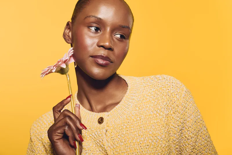 Portrait of a thoughtful black woman with dark skin holding a pink flower near her face. She wears a yellow knitted sweater against a bright yellow background. Concept of beauty and contemplation.