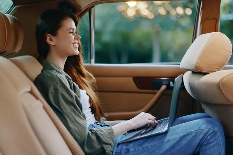 Young woman joyfully working on a laptop in a car, dressed casually in a green jacket and jeans, enjoying a sunny day with a blurred green background.