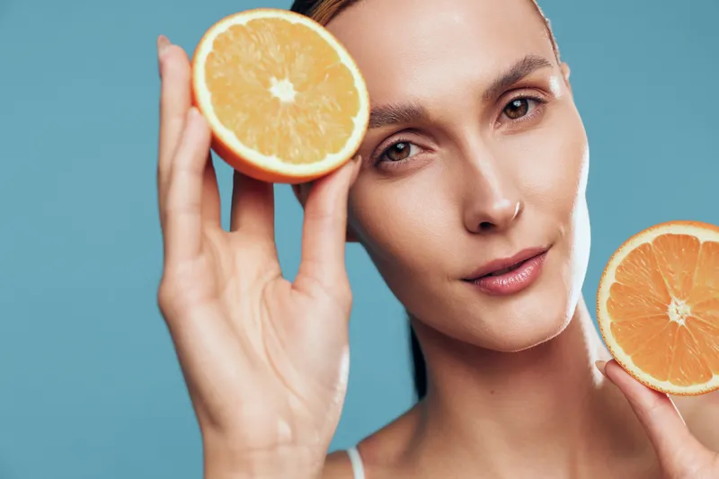 A young woman holds two halves of a fresh orange close to her face against a blue background. The image highlights healthy lifestyle, natural beauty, and fresh fruit concepts.