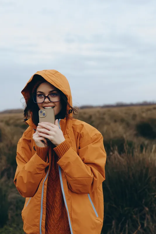 Woman lifestyle portrait outdoors in orange hooded jacket and glasses holding a smartphone in a field under cloudy sky Soft natural light suggests communication and calm everyday life