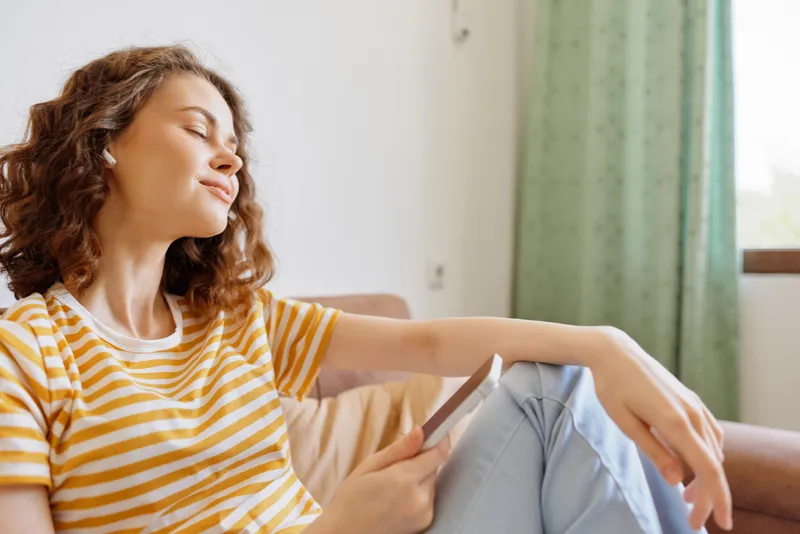 Woman relaxing at home on a couch, reading a book with a content smile, casual lifestyle scene, soft natural light, cozy interior, peaceful domestic moment.