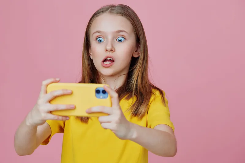 Surprised young girl in a yellow shirt looking at her phone against a pink background, expressing emotions through her facial expression