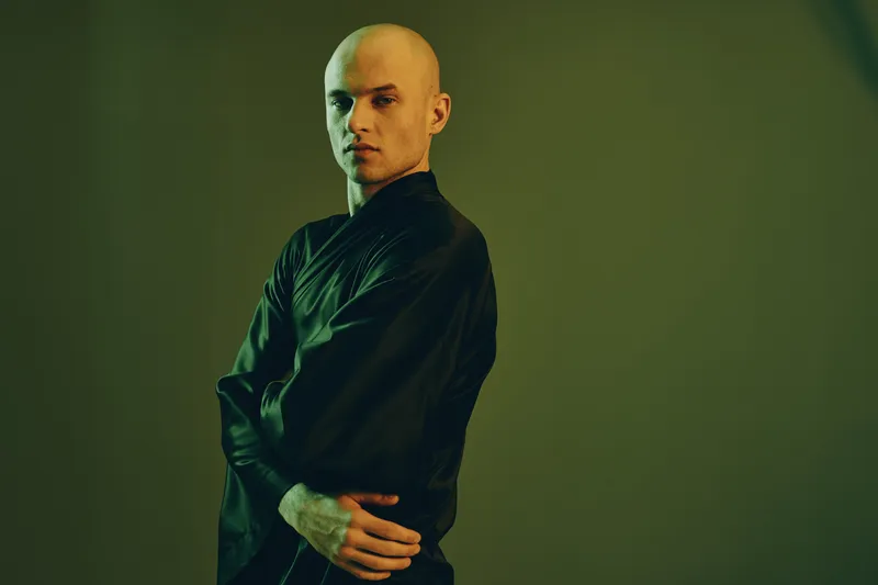 Confident man in black attire poses against a softly lit background, showcasing a modern aesthetic in muted tones. Emphasis on style and expression in composition.