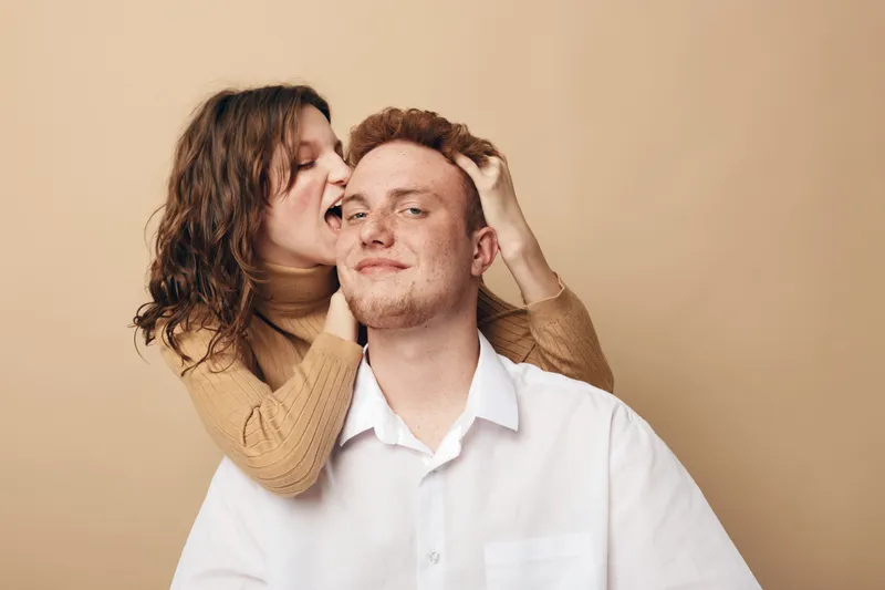 Young couple with red hair and curly hair playfully interact in beige studio. Woman bites man’s ear gently, both wearing casual clothes. Concept of love, fun, and intimacy.