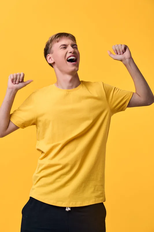 Energetic man celebrating in a bright yellow shirt, arms raised in joy and triumph, conveying excitement, happiness, and a festive, upbeat mood in a clean studio setting