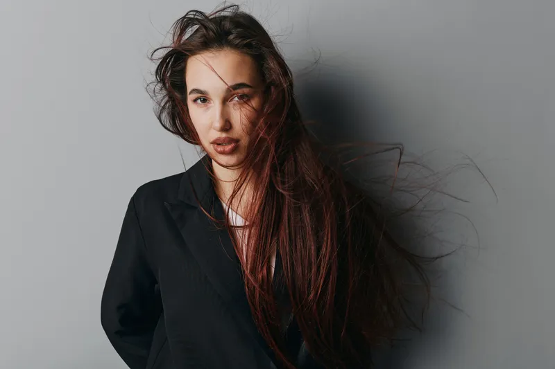 Woman with long hair and black jacket posing against gray background, looking at camera with serious expression, natural makeup and loose hairstyle in studio portrait.