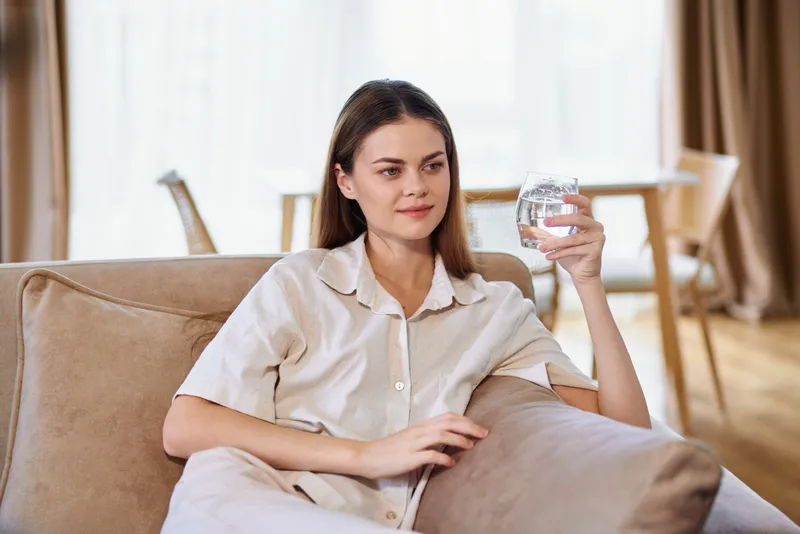 Woman on sofa holding a glass of water at home, relaxed and casual in light pajamas, portrait of comfort in a bright livingroom with soft cushions and natural window light.