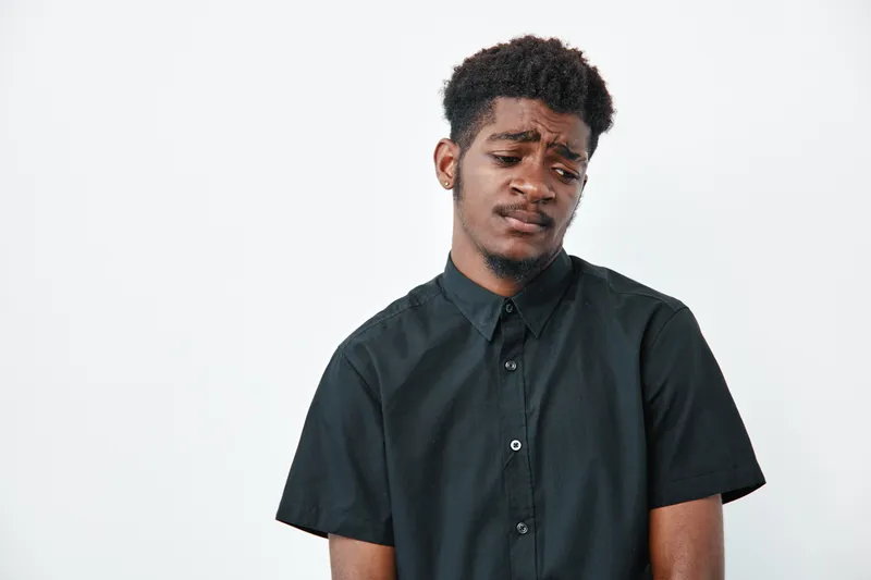 Young African American man with dark skin wearing black shirt looking down with sad expression isolated on white background. Portrait, emotion and casual style.