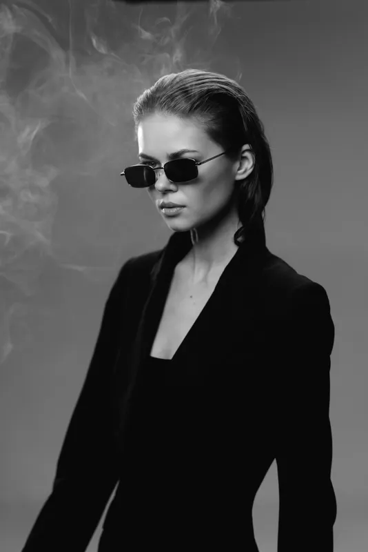 Black and white portrait of confident woman wearing sunglasses and stylish black outfit with smoke background