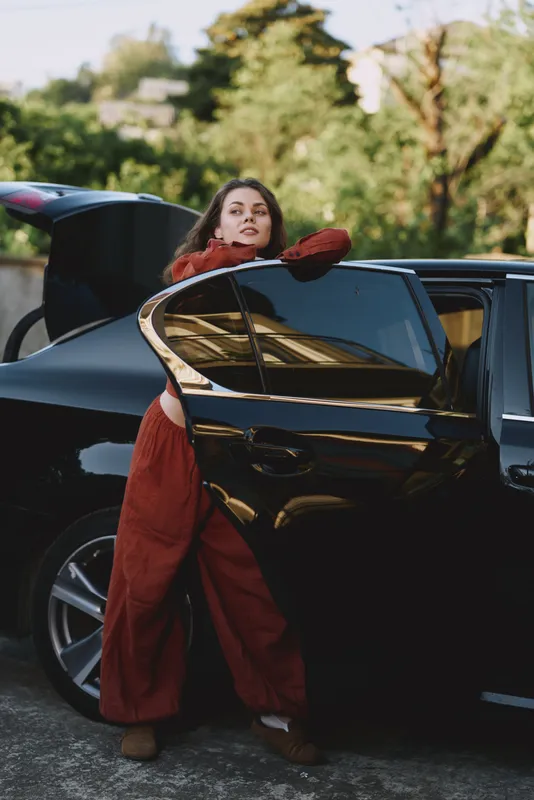 Cheerful woman outdoors by a sleek car, resting against the door with a relaxed pose, red outfit, natural light, city park background, candid lifestyle moment