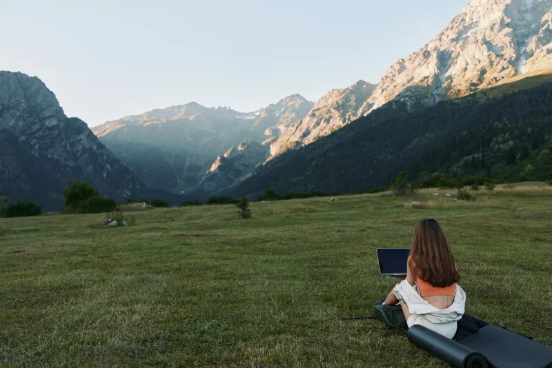 A person sits on a grassy hillside with a laptop, surrounded by rugged mountains and clear sky. They work outdoors, appearing focused and relaxed, creating a calm and productive atmosphere.