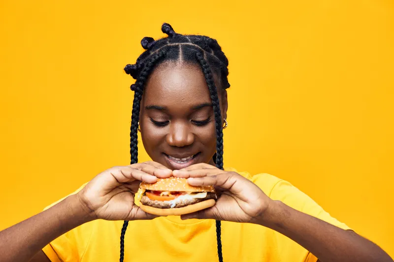 Joyful young girl in a yellow shirt holding a delicious hamburger with a bright yellow background, showcasing happiness and a fun food experience