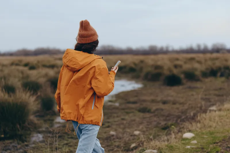 Woman outdoor walking lifestyle on countryside field path, wearing orange jacket and knit hat, holding phone, looking ahead, soft daylight, calm casual travel mood, in nature landscape view