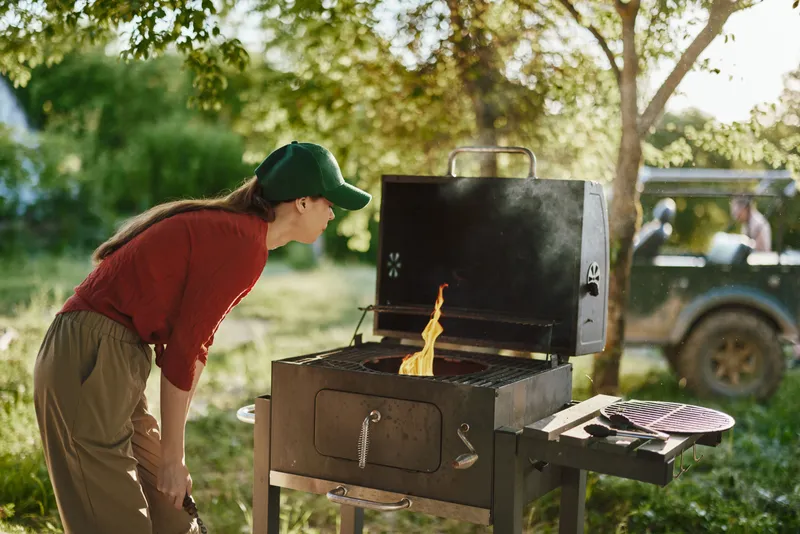 Young woman grilling outdoors, smiling, wearing a green cap and red shirt, enjoying a summer barbecue in a lush green garden setting