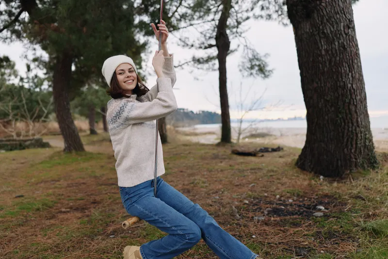 woman on a swing in outdoors nature forest, wearing a sweater and scarf, enjoying a joyful moment among trees and grass in a peaceful landscape