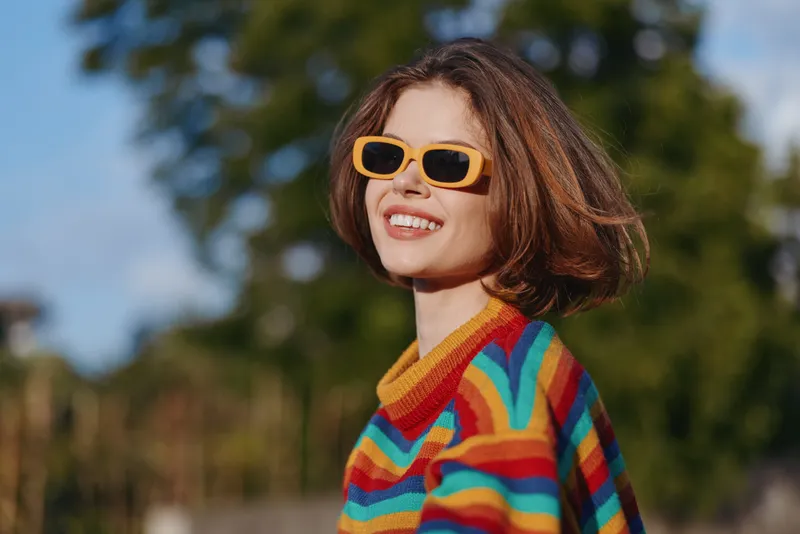 Woman smiling in colorful striped sweater and yellow sunglasses, short hair portrait outdoors in sunny park setting. Casual fashion, confident expression for lifestyle and travel use.