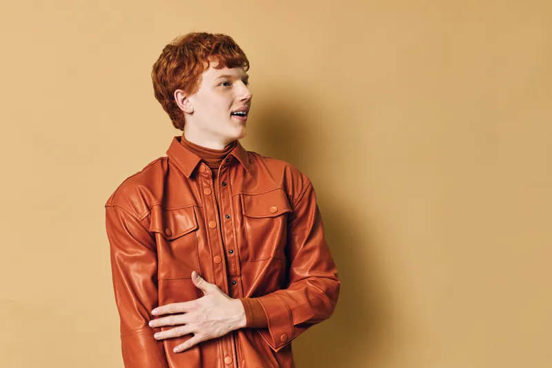Young man with red hair and freckles wearing a brown leather jacket posing against beige background looking to the side with natural expression and casual style.