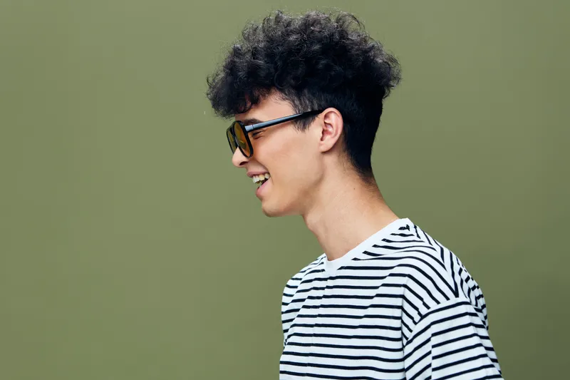 Young man with curly hair wearing sunglasses and striped t-shirt smiles happily against a soft green background, displaying casual and carefree style.