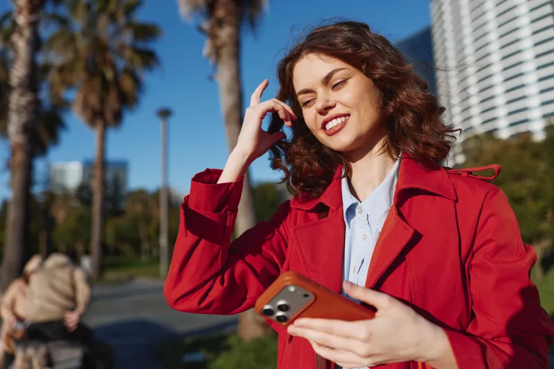 Young woman with curly hair wears red coat and smiles while holding smartphone outdoors on a sunny day in an urban park setting.