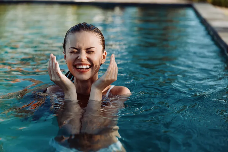 woman pool water smiling happiness summer enjoying sunny afternoon in the pool with hands near face, playful moment, relaxed lifestyle scene by water, candid joy and refreshment