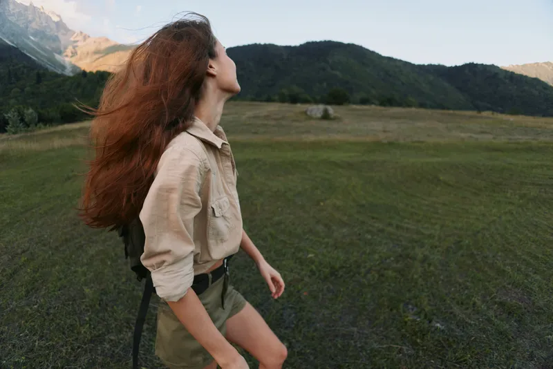 Young woman with long red hair standing in a green field looking up at the sky. Casual outfit with beige shirt and shorts. Concept of lifestyle and nature.