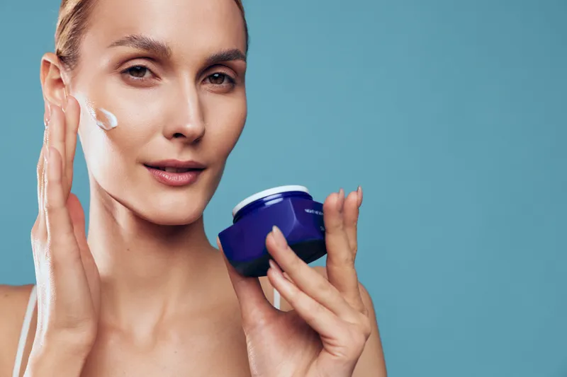 Young woman with brunette hair applies cream to her cheek in a studio scene, demonstrating a skincare routine with calm focus, soft lighting, and a clean blue background to emphasize skin care