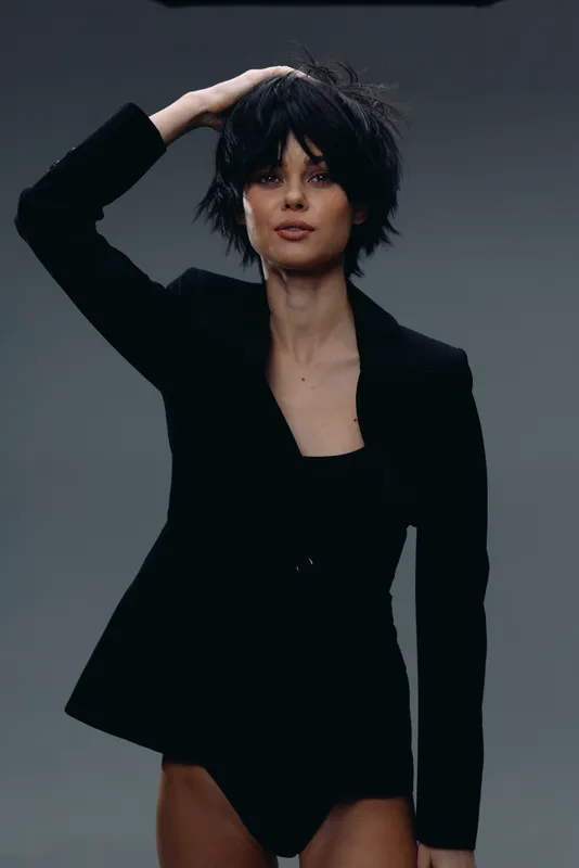 Stylish woman with short black hair wearing a black blazer posing confidently against a gray background.
