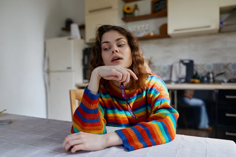Young woman with curly brown hair sits at a bright kitchen table wearing a colorful striped sweater, relaxed and focused, conveying a cozy home lifestyle