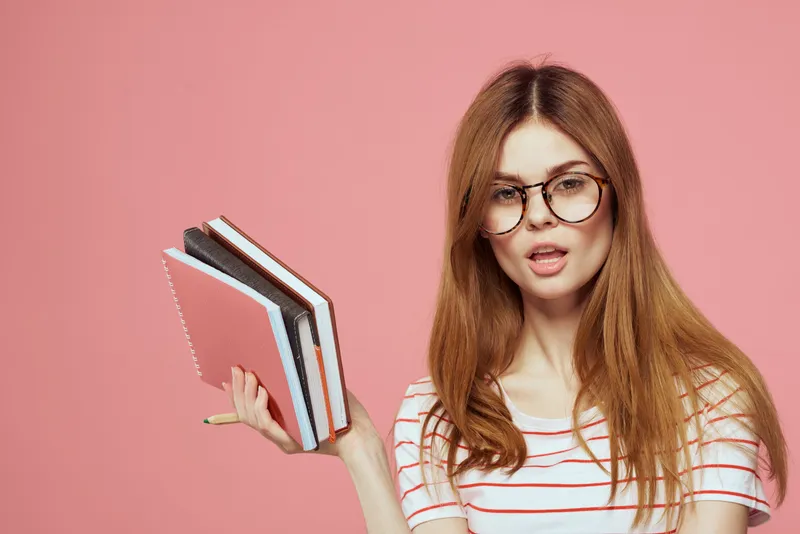 Красивая студентка держит книги, жестикулирует руками на розовом фоне