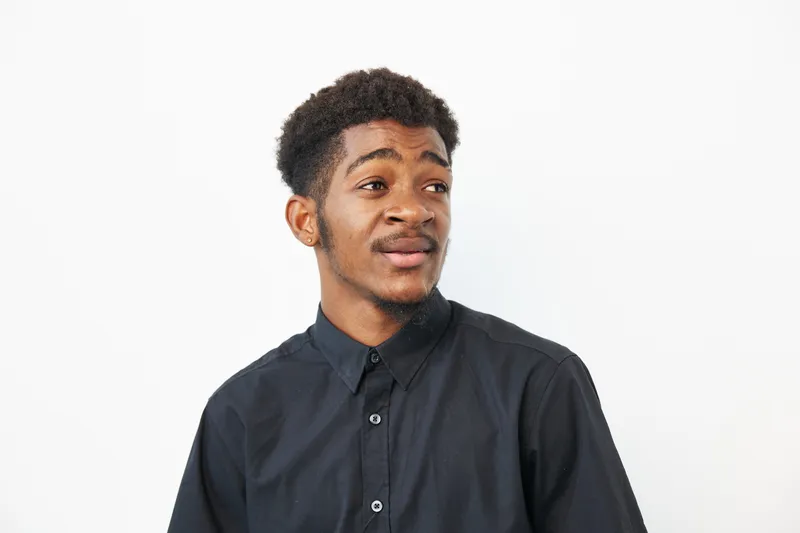 A thoughtful black man with an expressive face, wearing a black shirt against a clean white background, radiating confidence and curiosity in his gaze.