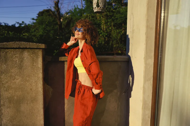 Cheerful woman outdoors in orange suit and yellow top, leaning on a terrace railing, sunglasses on, sunny moment reflects casual lifestyle, stylish fashion, relaxed posture