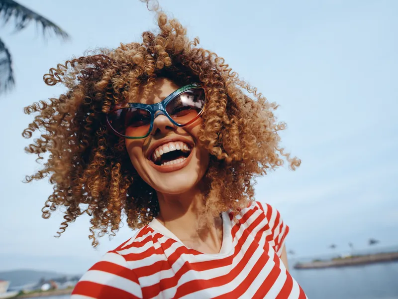 Vibrant portrait of a joyful woman with curly hair wearing a red and white striped shirt and oversized sunglasses, captured outdoors under a clear blue sky, smiling boldly.