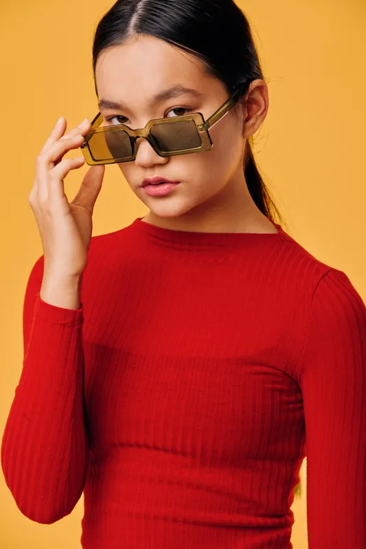 Young woman in red sweater adjusting brown sunglasses while posing against bright orange background in studio. Fashion portrait with confident expression and casual style for modern look.