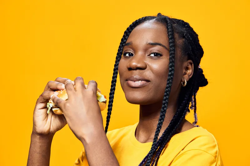 A young Black woman joyfully enjoying a healthy burger, showcasing vibrant creativity against a bright yellow background, perfect for food related content