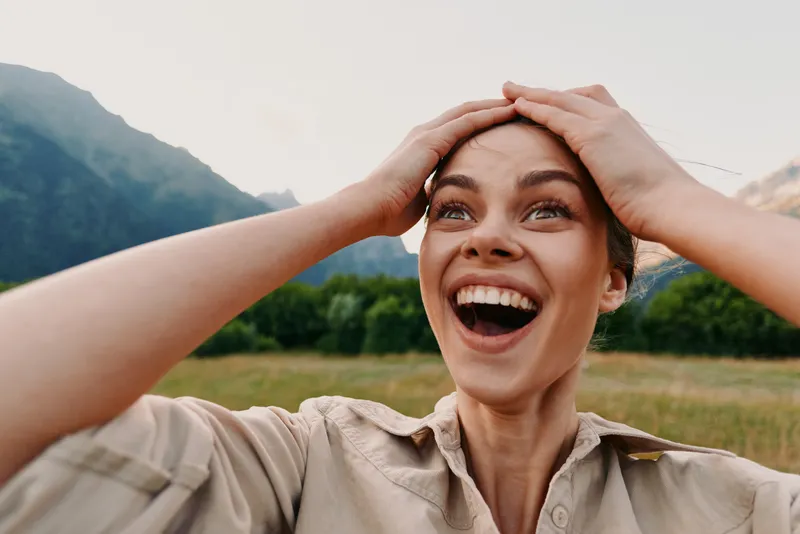 Young woman with short hair laughs joyfully while holding her head in a natural outdoor setting with mountains and greenery in the background.