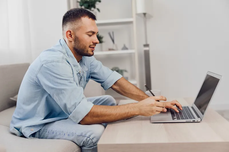 Focused man working on a laptop while sitting on a couch, wearing casual attire, showcasing productivity, modern workspace, and creative vibe.