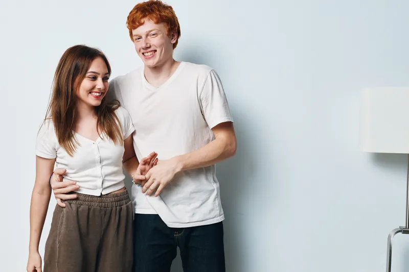 Couple, young, smiling and standing with casual clothes near white wall indoors. Man and woman holding hands in relaxed pose with friendly expressions and soft natural light.