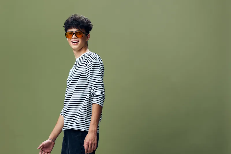 Smiling young man wearing striped shirt and orange sunglasses stands against green background, radiating casual style and joyful mood in studio setting.
