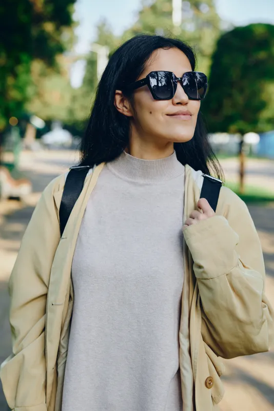 Casual fashion in the park: a woman wearing oversized sunglasses and a beige coat with a soft knit sweater, carrying a backpack, enjoying sunny outdoor scenery.
