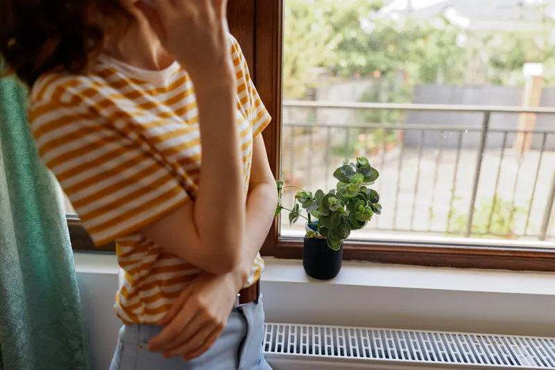 Woman at home lifestyle by a window, plant on the sill, talking on a phone, casual striped shirt, relaxed mood in a cozy living space, intimate everyday scene