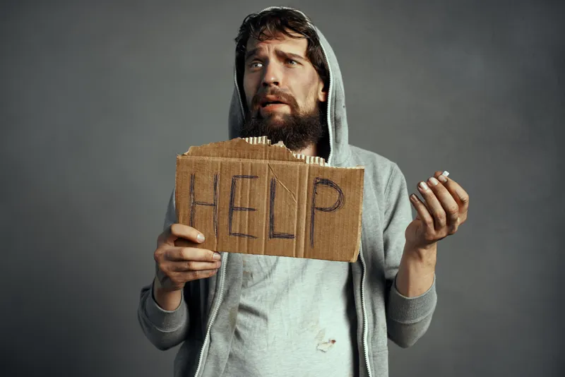 Бездомный мужчина с бородой держит табличку с надписью «Help» на улице