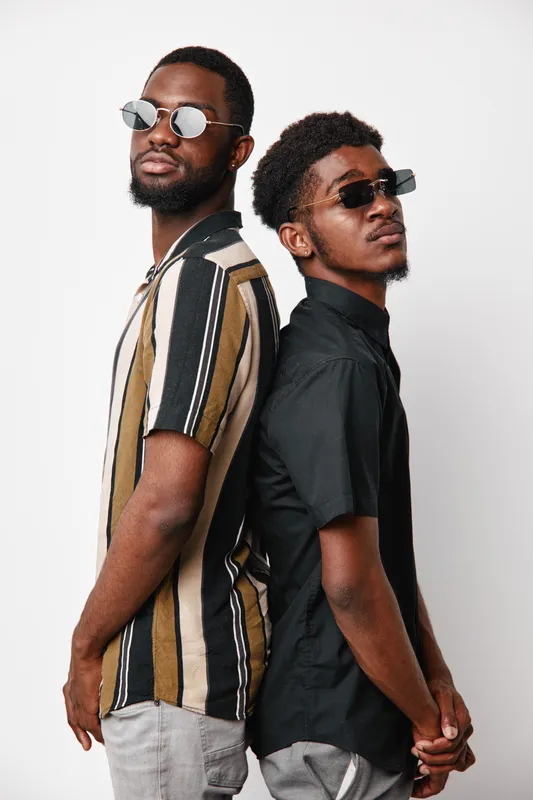 Two African American men with dark skin wearing sunglasses and casual shirts standing back to back against white background. Young adults with confident expressions and modern style.