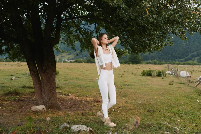 Young woman in white outfit stands relaxed in a sunlit countryside, arms behind her head, basking under a large tree with grazing sheep in the background.