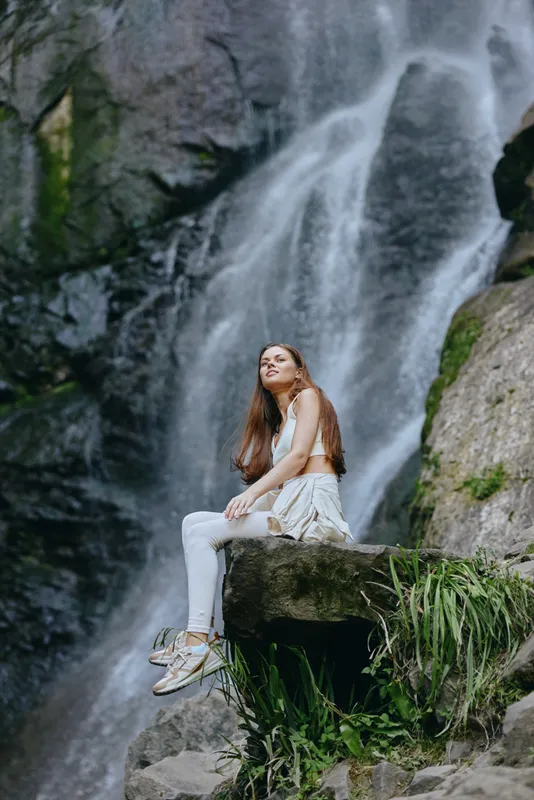 young woman sitting on rock near waterfall, wearing white activewear, peaceful outdoor nature scene, greenery and flowing water background