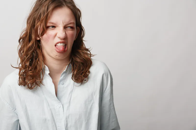 Young woman with curly hair in white shirt makes disgusted face, sticking out tongue. Isolated on light background. Concept of dislike, rejection, and negative emotions in casual style.