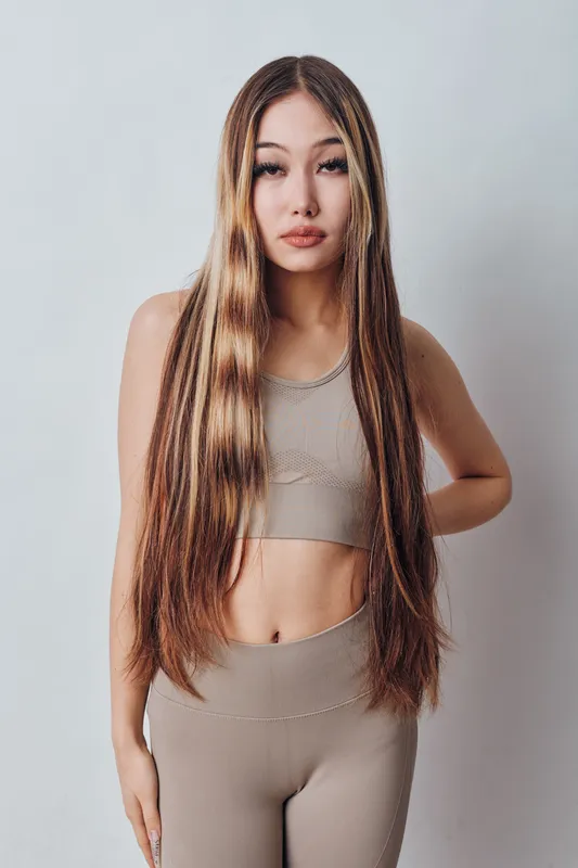 Young woman with long straight hair standing and posing in beige outfit against plain background. Fashion model showing confident expression with relaxed posture and natural look.