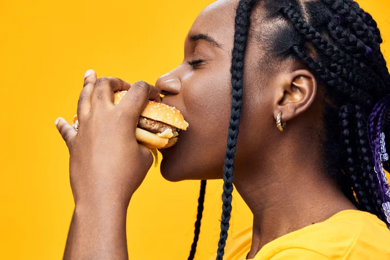 Young woman enjoying a delicious burger against a vibrant yellow background, exuding happiness and satisfaction This image captures a moment of joyful eating with a focus on fast food